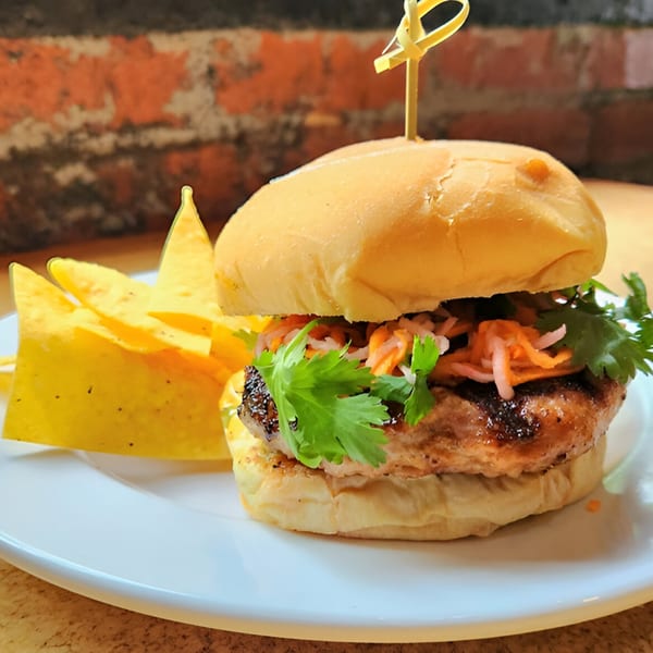 A grilled burger with pickled vegetables and cilantro, served with tortilla chips on a white plate.